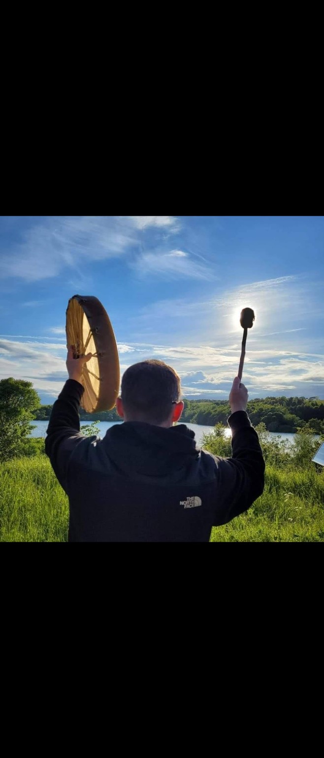 A person raising a frame drum beside a loch